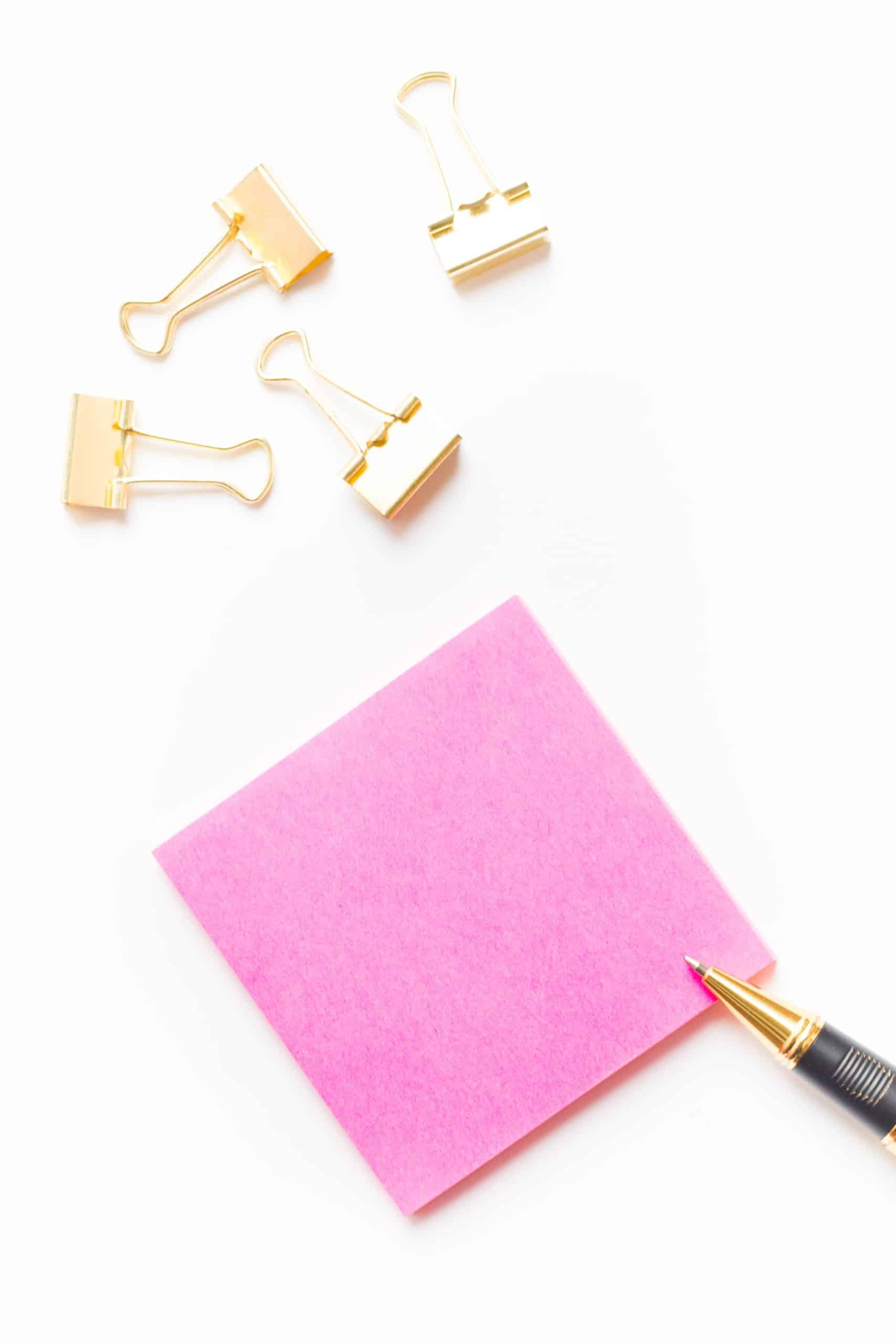 A pink sticky note with the tip of a black and gold pen touching its bottom right corner. Four gold binder clips are placed nearby on a white background, perfect for creating an editable template that caters to extended use.