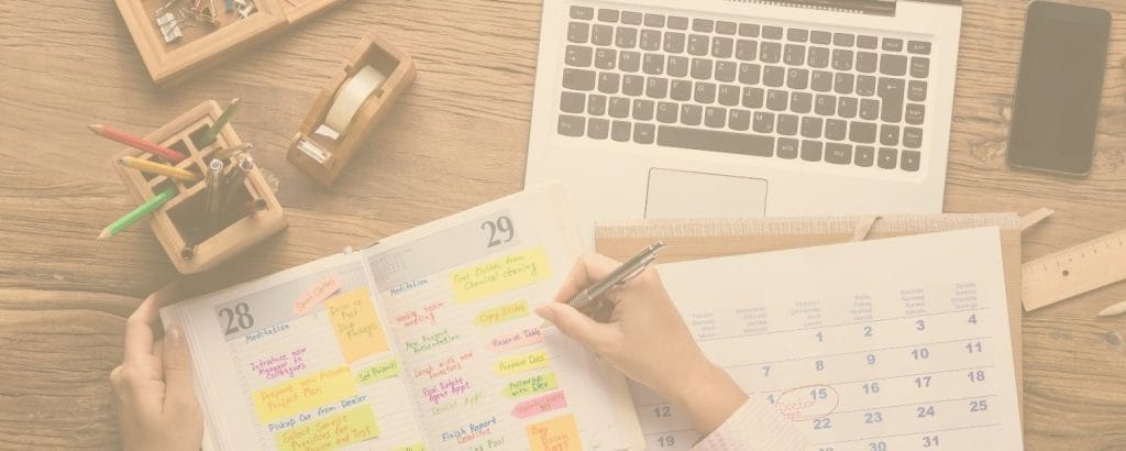 A person writes in a colorful planner next to a calendar, with a laptop, smartphone, wooden organizer, and stationery on a wooden desk.